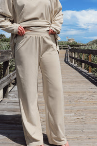 A woman wearing a matching beige lounge set consisting of a long-sleeve top and wide-leg pants, standing on a wooden boardwalk outdoors.