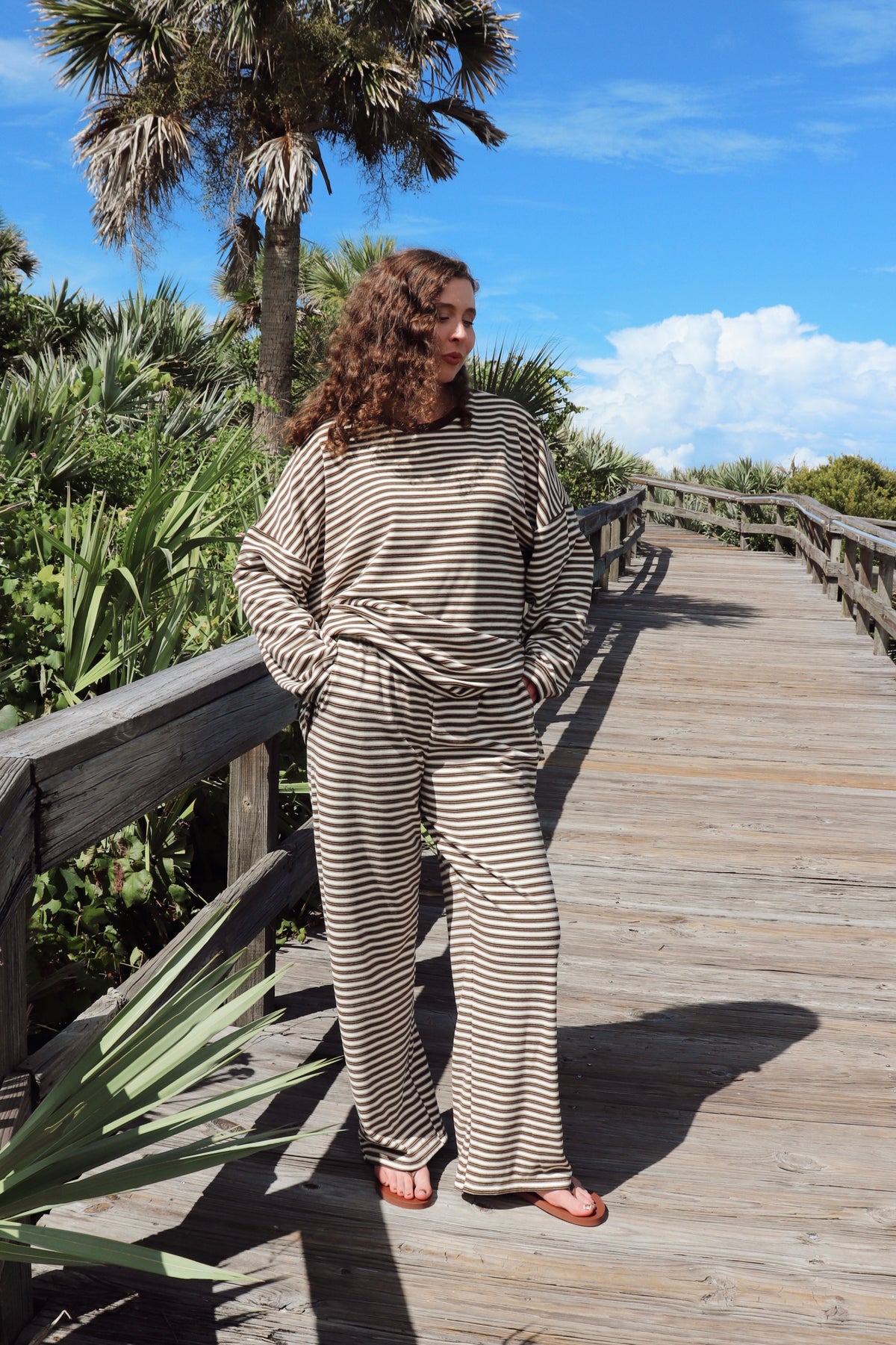 A woman wearing a beige and brown striped lounge set stands on a wooden boardwalk surrounded by green foliage and palm trees under a blue sky.
