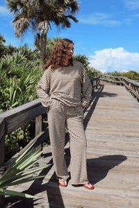 A woman wearing a beige and brown striped lounge set stands on a wooden boardwalk surrounded by green foliage and palm trees under a blue sky.