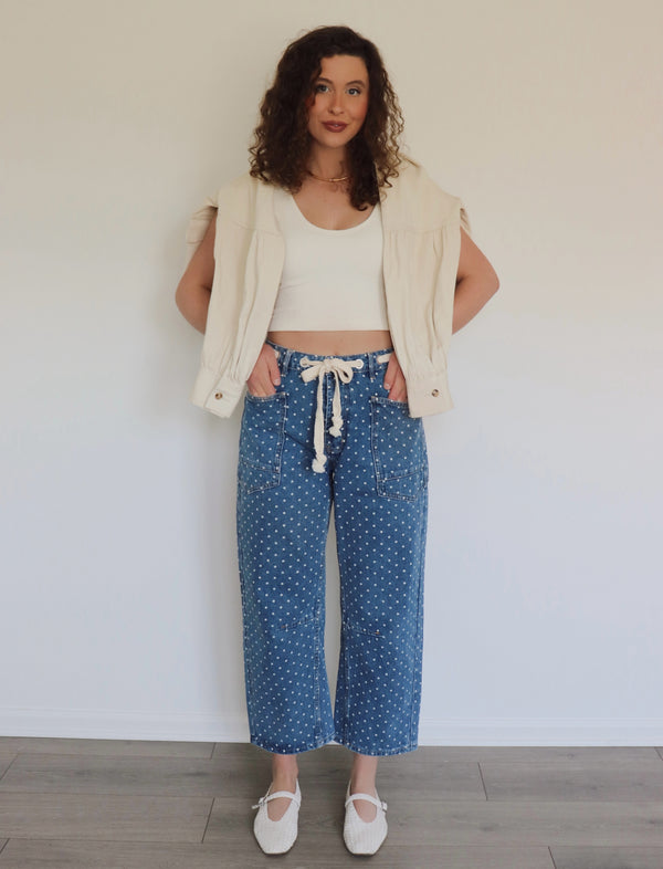 woman wearing white tank top and blue jeans with white polka dots on them against a white wall
