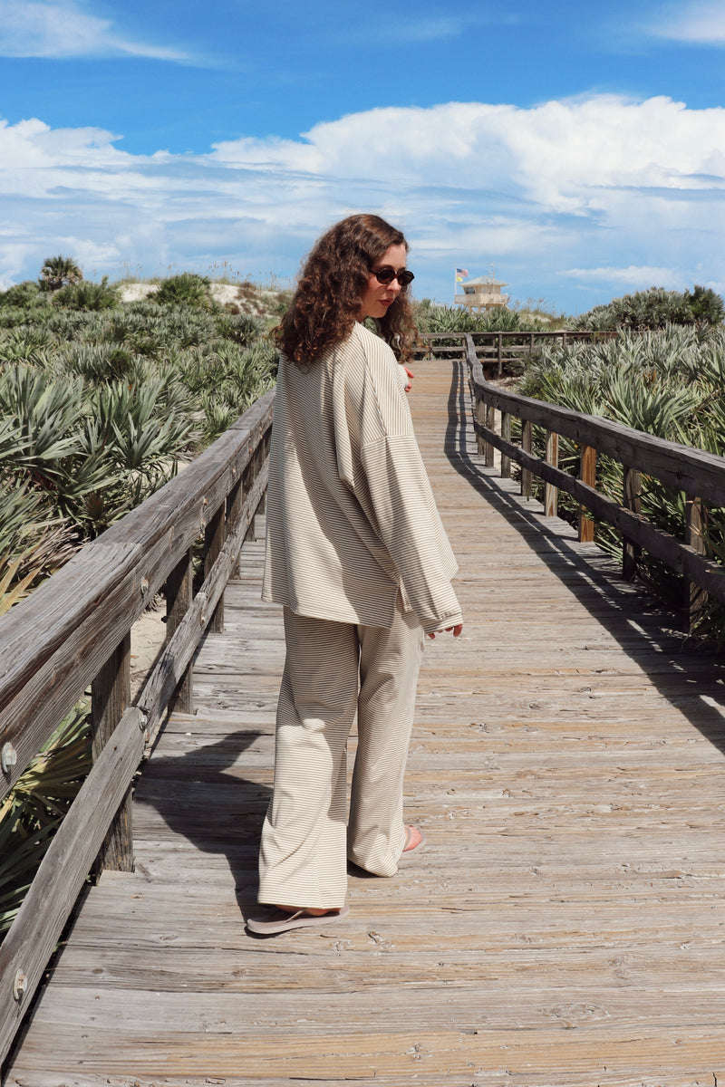 A woman wearing a matching beige lounge set consisting of a long-sleeve top and wide-leg pants, standing on a wooden boardwalk outdoors.