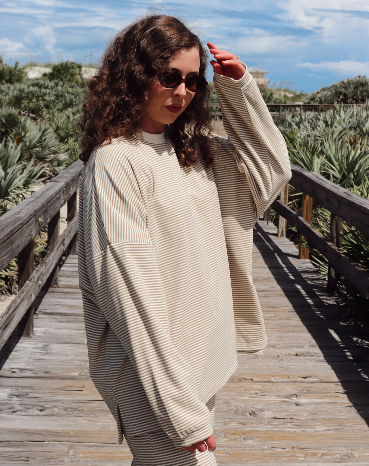 A woman wearing a matching beige lounge set consisting of a long-sleeve top and wide-leg pants, standing on a wooden boardwalk outdoors.