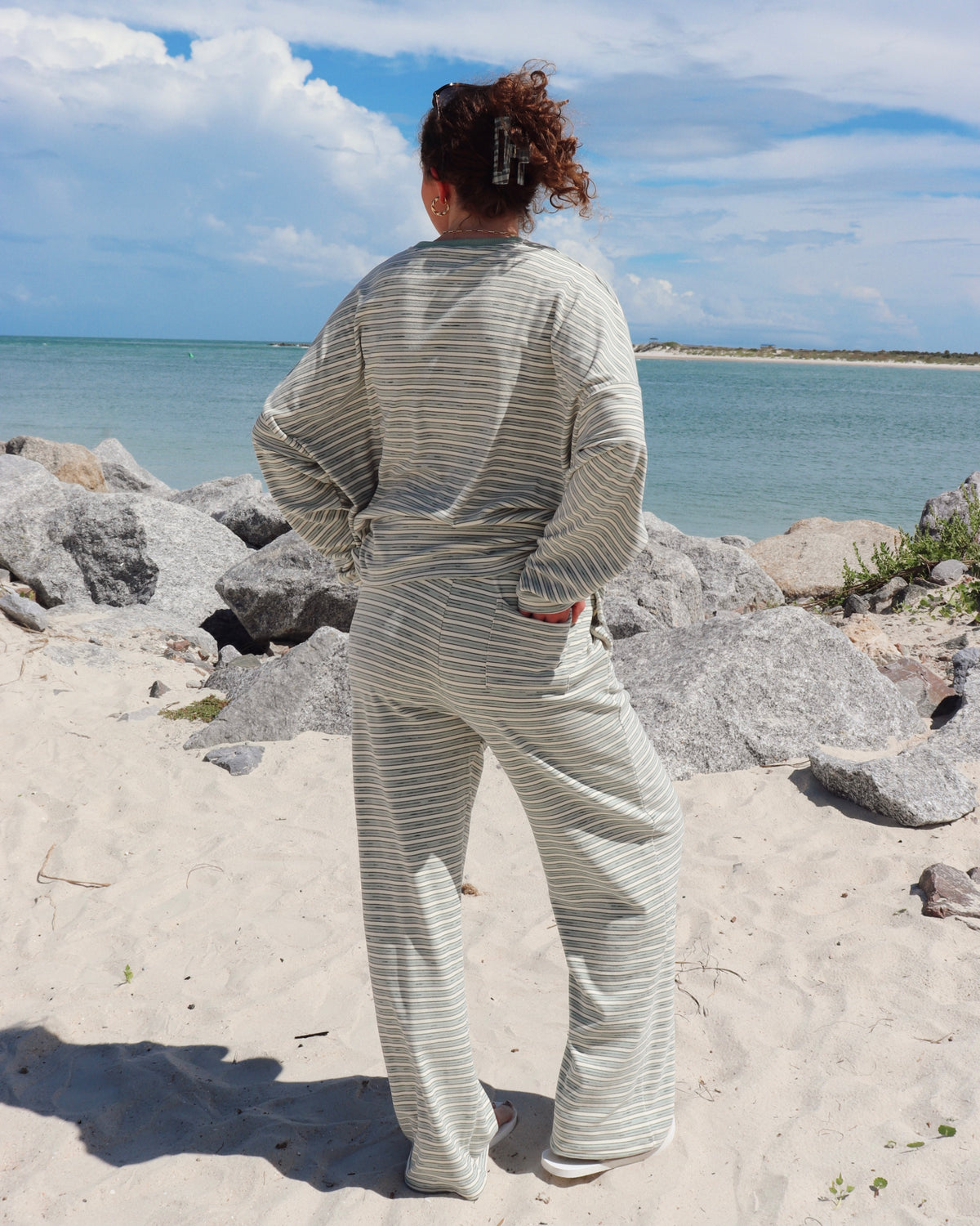 A woman stands on a sandy beach with rocks and the ocean in the background, viewed from behind. She is wearing a matching long-sleeve top and wide-leg pants set in a light mint green and white stripe pattern.