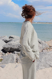A woman with curly brown hair stands on a rocky beach facing the ocean, wearing a light mint green and white striped long-sleeve top and matching bottoms.