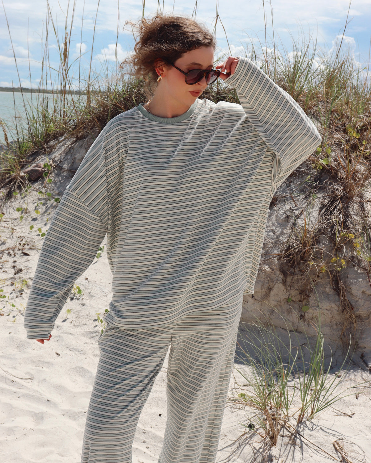 A woman wearing a matching two-piece lounge set with thin light mint green and white stripes stands on a sandy beach with dune grass and water in the background.