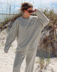 A woman wearing a matching two-piece lounge set with thin light mint green and white stripes stands on a sandy beach with dune grass and water in the background.