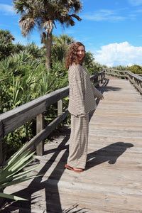 A woman wearing a beige and brown striped lounge set stands on a wooden boardwalk surrounded by green foliage and palm trees under a blue sky.