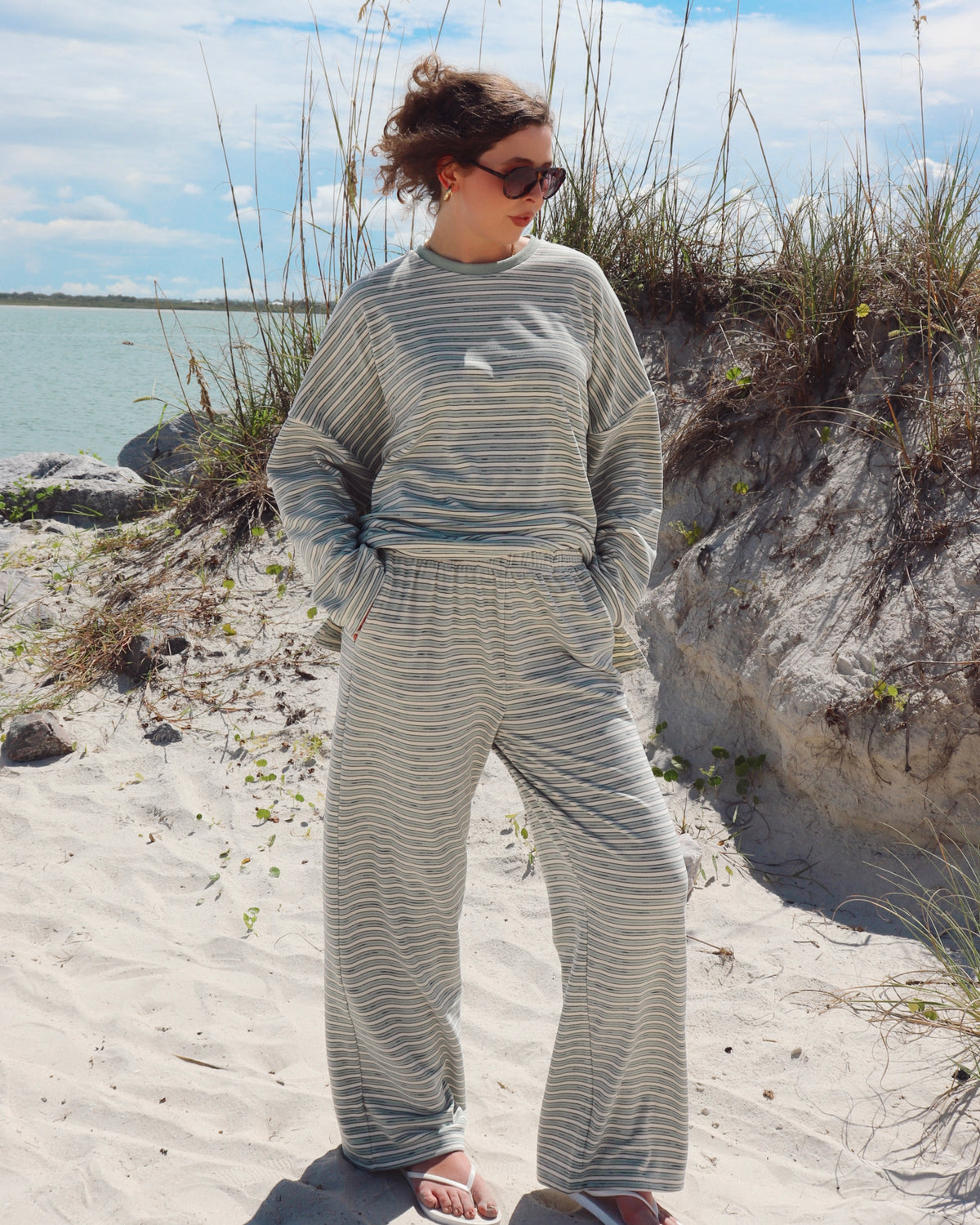 A woman wearing a matching two-piece lounge set with thin light mint green and white stripes stands on a sandy beach with dune grass and water in the background.