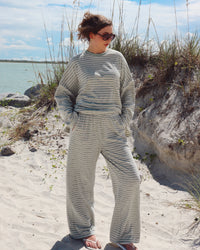 A woman wearing a matching two-piece lounge set with thin light mint green and white stripes stands on a sandy beach with dune grass and water in the background.