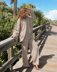 A woman wearing a beige and brown striped lounge set stands on a wooden boardwalk surrounded by green foliage and palm trees under a blue sky.