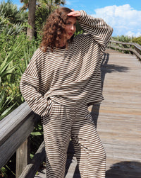 A woman wearing a beige and brown striped lounge set stands on a wooden boardwalk surrounded by green foliage and palm trees under a blue sky.