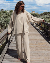 A woman wearing a matching beige lounge set consisting of a long-sleeve top and wide-leg pants, standing on a wooden boardwalk outdoors.