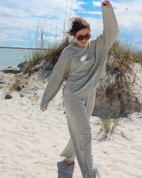 A woman wearing a matching two-piece lounge set with thin light mint green and white stripes stands on a sandy beach with dune grass and water in the background.