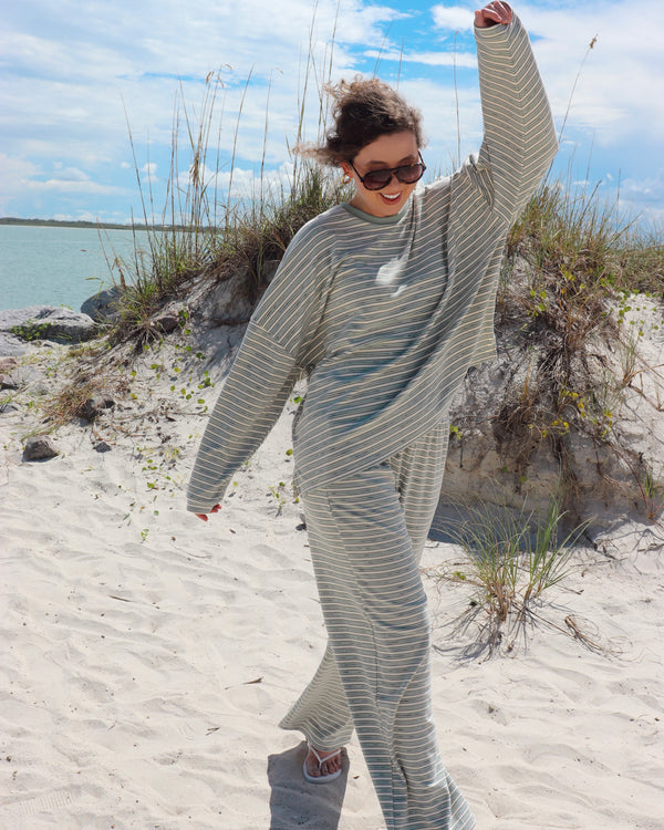A woman wearing a matching two-piece lounge set with thin light mint green and white stripes stands on a sandy beach with dune grass and water in the background.