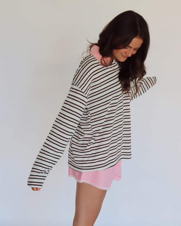 A woman modeling a brown, pink and white horizontally striped long-sleeve polo shirt with a pink collar, paired with light pink shorts, against a plain white background.