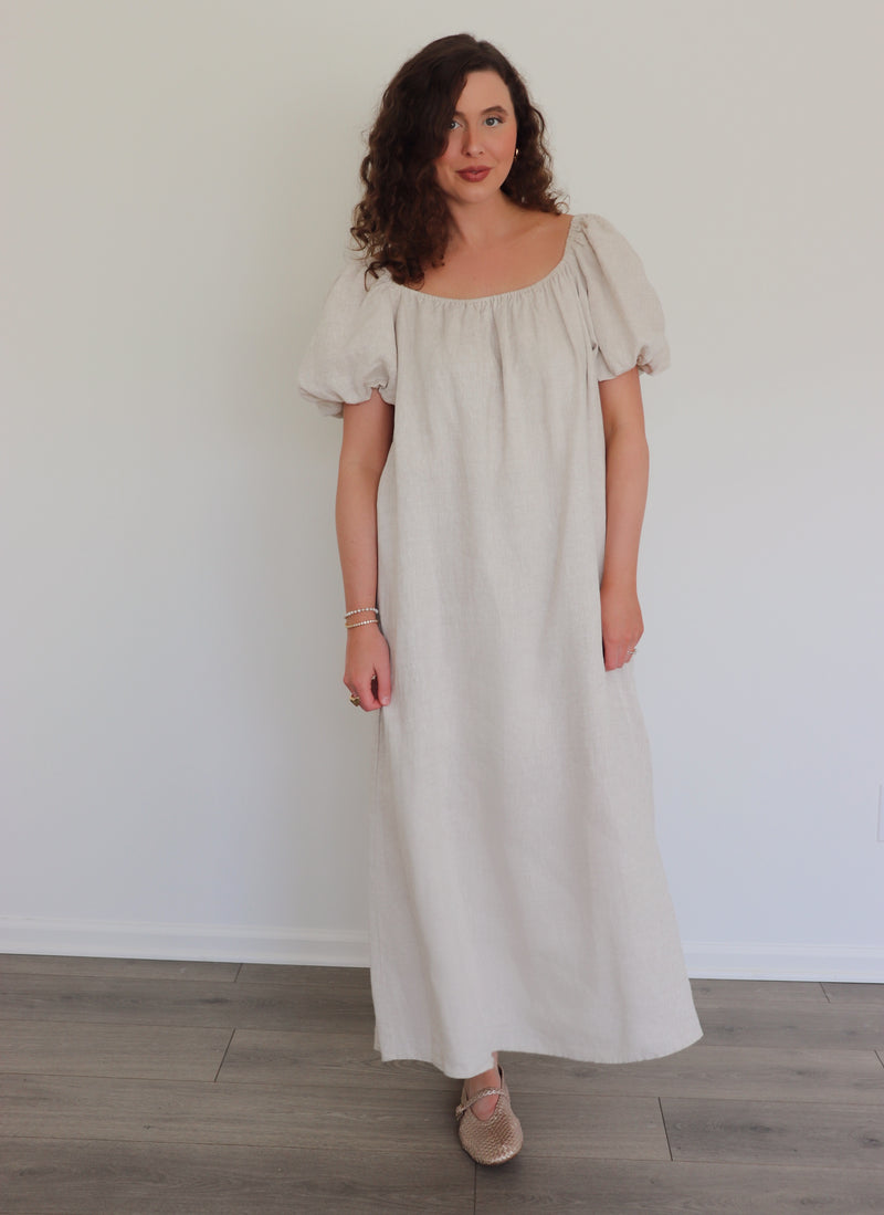 Woman wearing a long beige linen dress against a white wall