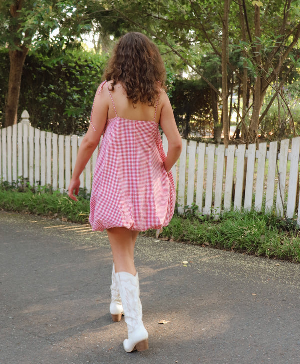 Woman in a red checkered dress and white boots standing on a path with trees and a white fence in the background.