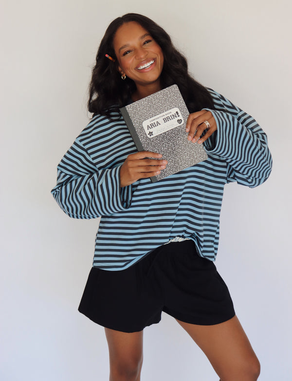 A woman smiling and holding a notebook with a white label that reads 'Aria+ Brin'. She is wearing a blue and navy horizontally striped long-sleeve top and shorts, standing against a plain white background.