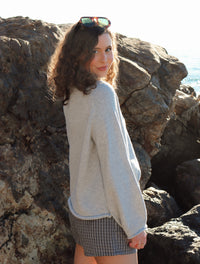 Woman outdoors by a rocky coastline, wearing a gray sweater and black shorts. 