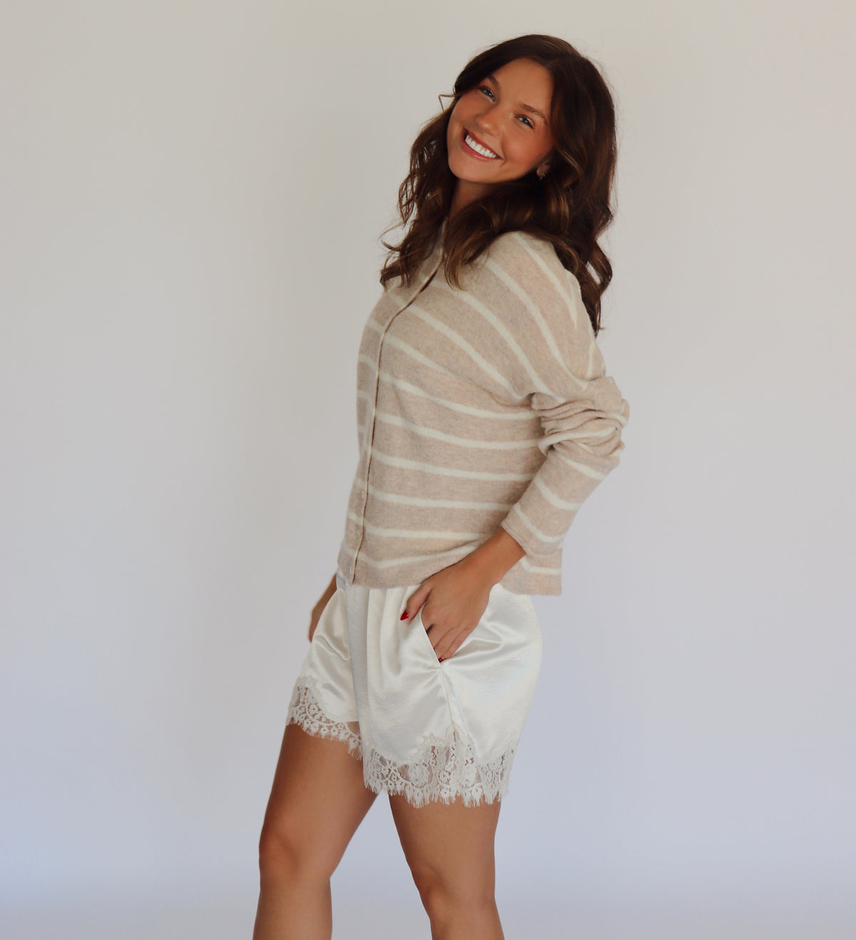 A woman modeling a beige and white striped knit long-sleeve top paired with white satin shorts featuring lace trim, standing against a plain white studio background.