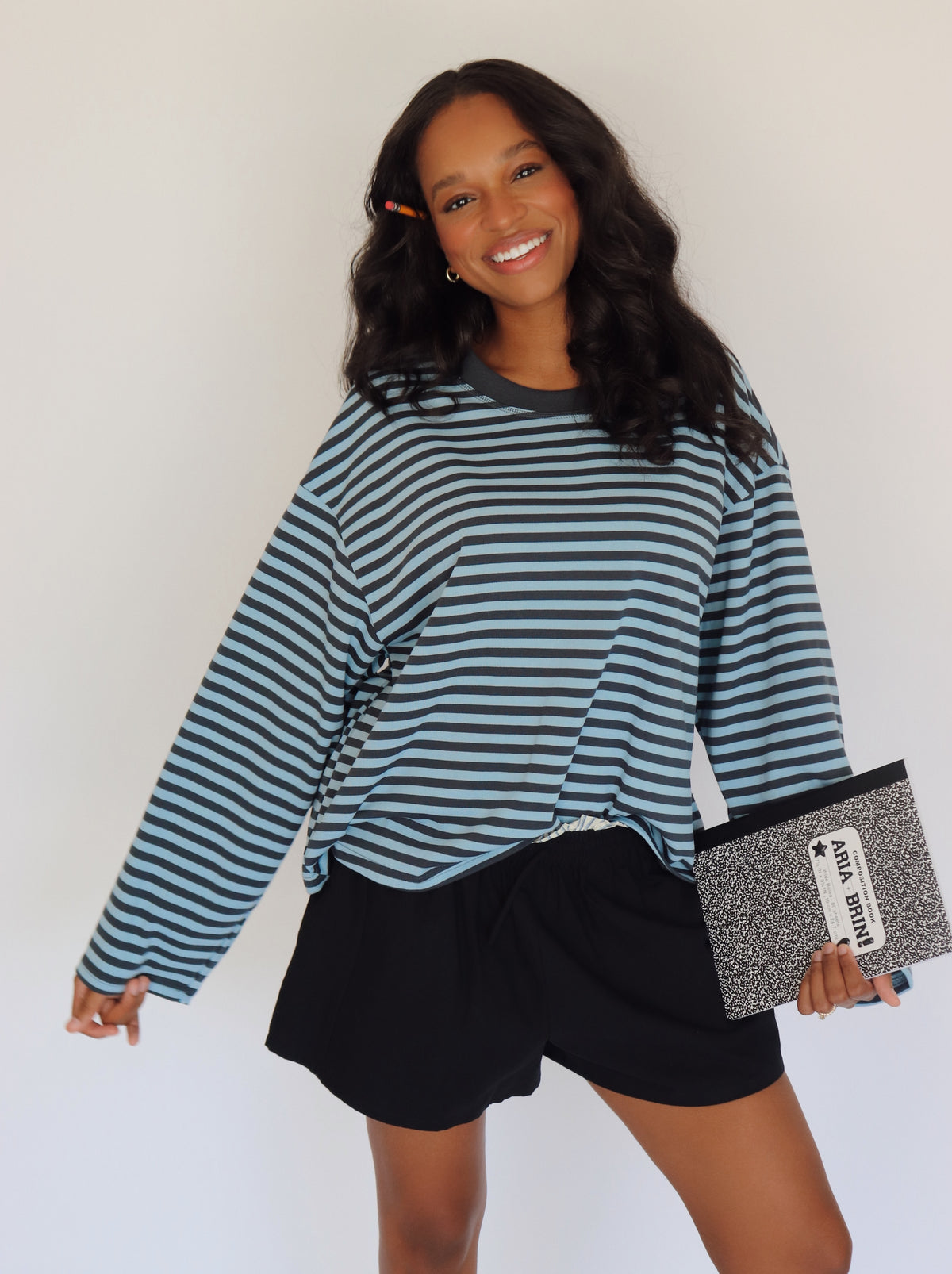 A woman modeling a long-sleeve blue striped top with shorts, holding a black and white notebook bag against a plain white background.