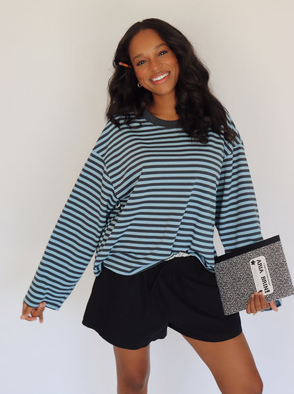A woman modeling a long-sleeve blue striped top with shorts, holding a black and white notebook bag against a plain white background.