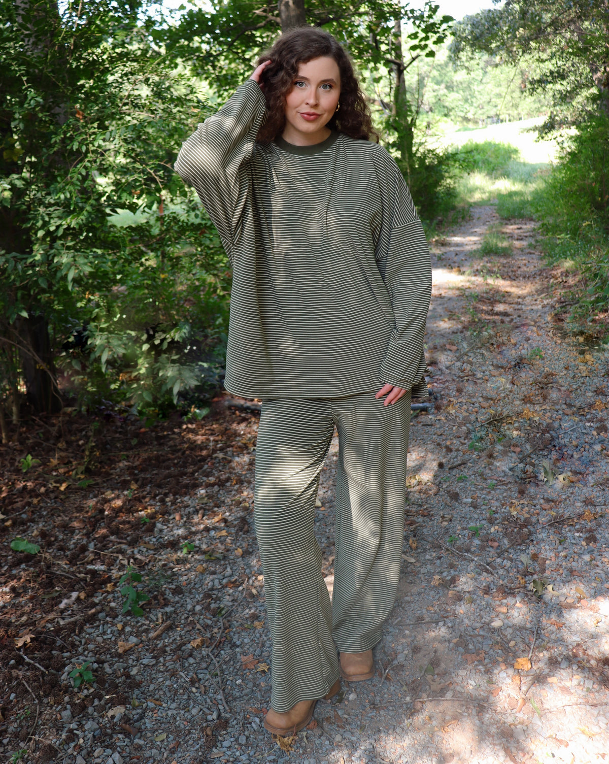 A woman wearing a matching olive green striped lounge set stands outdoors in a wooded area with greenery and. a gravel road.