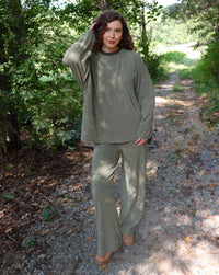 A woman wearing a matching olive green striped lounge set stands outdoors in a wooded area with greenery and. a gravel road.