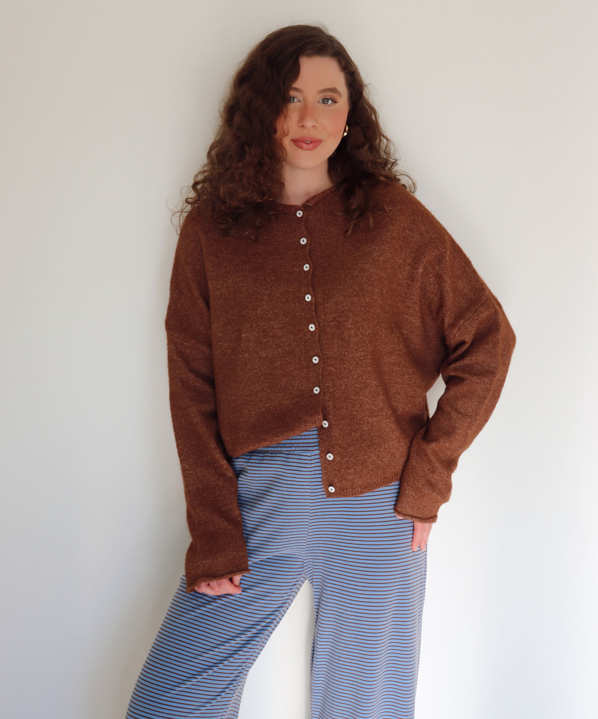 Woman wearing a brown cardigan over blue and brown striped pants against a plain background.