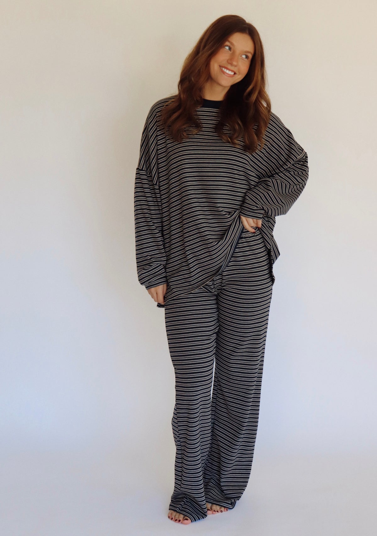 A woman modeling a matching two-piece loungewear set consisting of a long-sleeve top and wide-leg pants, both featuring a navy and white horizontal stripe pattern, set against a plain white studio background.