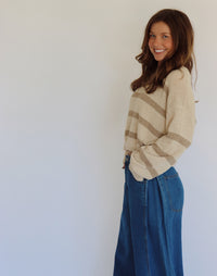 A woman wearing a taupe and tan striped knit sweater and high-waisted blue jeans, posing against a plain light gray wall.