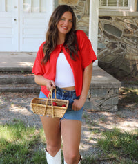 Woman in red top and denim shorts holding a basket near a porch.
