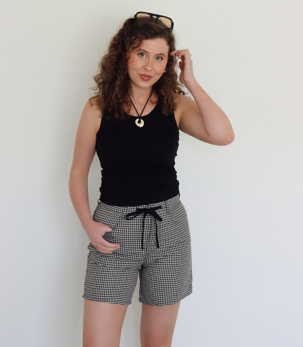 A woman with curly brown hair wearing a black tank top and black-and-cream gingham shorts, standing against a plain white wall.