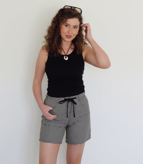 A woman with curly brown hair wearing a black tank top and black-and-cream gingham shorts, standing against a plain white wall.