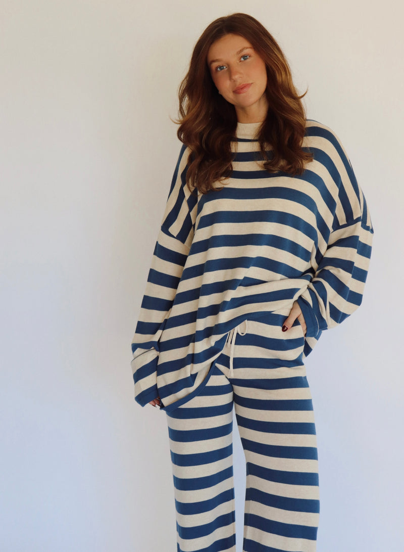 A woman wearing a matching two-piece loungewear set with blue and beige horizontal stripes, featuring a long-sleeve top and wide-leg pants, standing against a plain white background.
