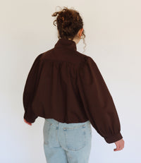 A person viewed from behind wearing a dark brown cropped blouse jacket with voluminous long sleeves and a gathered high neck, paired with light blue jeans against a plain white background.