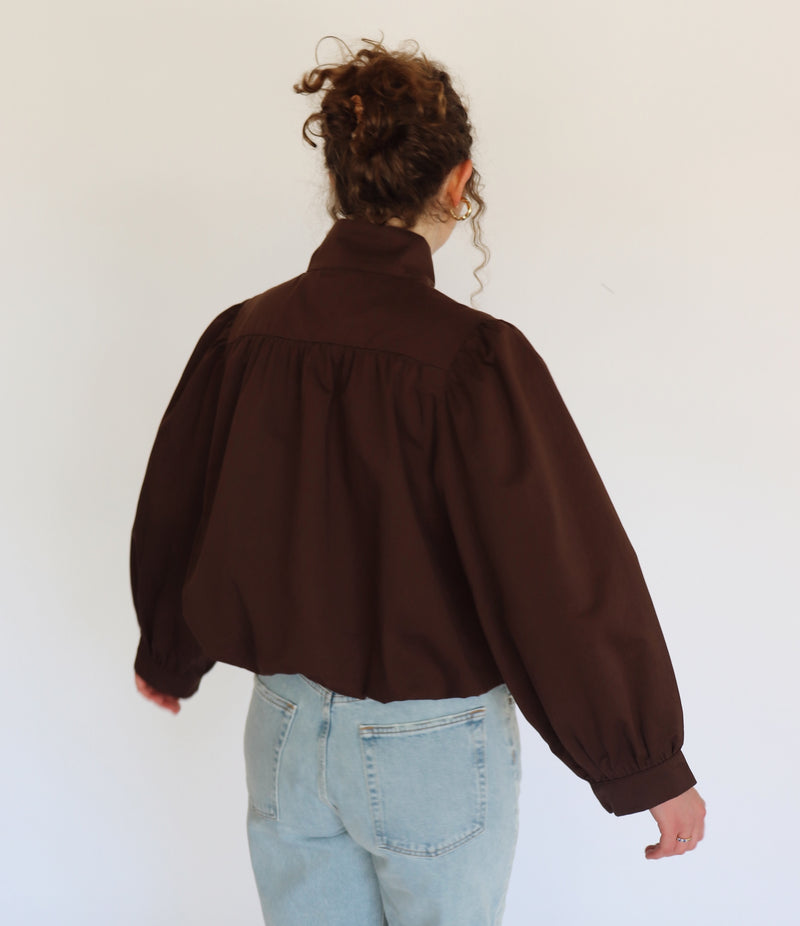 A person viewed from behind wearing a dark brown cropped blouse jacket with voluminous long sleeves and a gathered high neck, paired with light blue jeans against a plain white background.