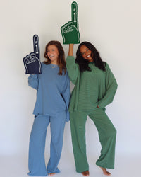 Two women modeling matching lounge sets, one in a dusty blue set and one in a dark green set, each holding a foam finger. The background is a clean white studio wall.