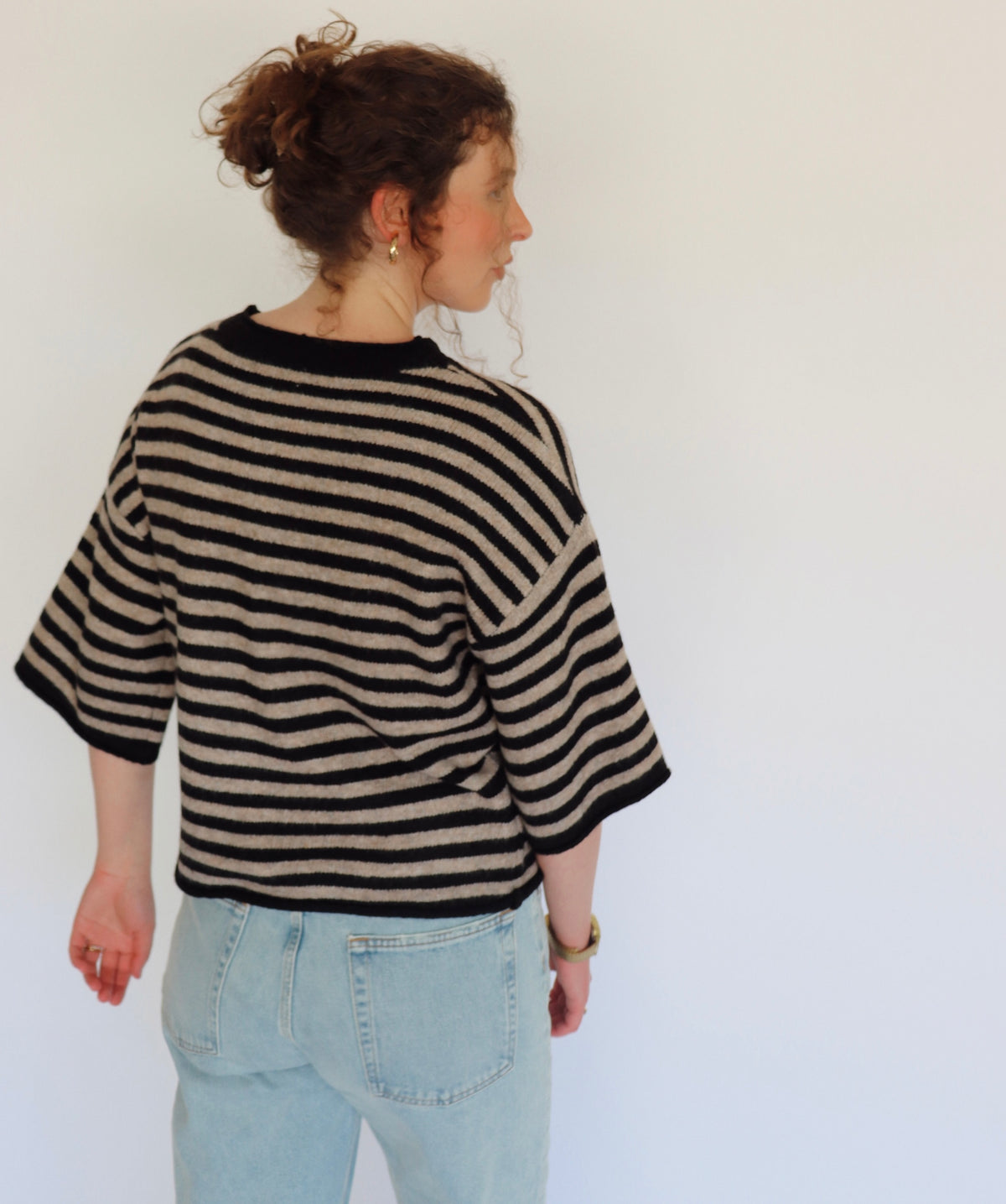 A woman shown from the back wearing a black and beige striped knit sweater with 3/4 sleeves and light blue jeans, standing against a plain white wall.