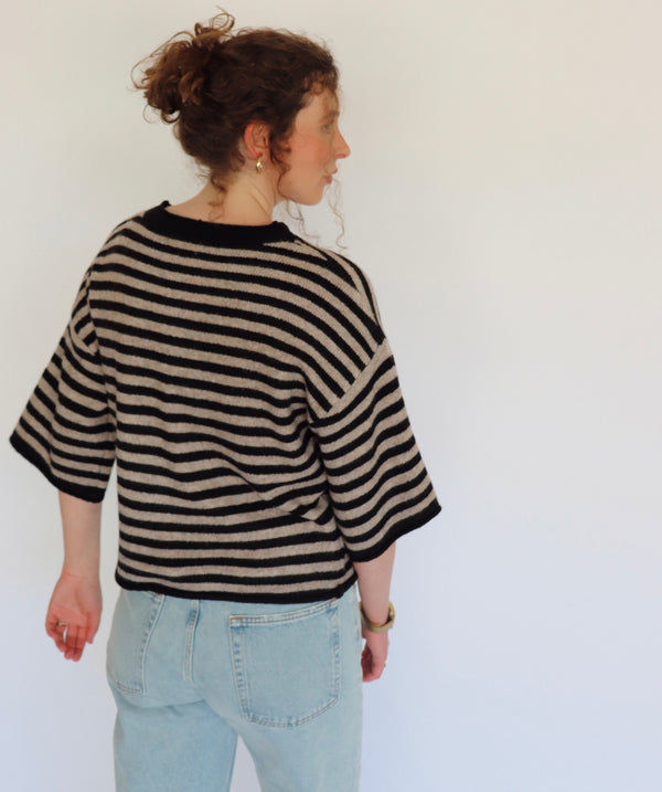 A woman shown from the back wearing a black and beige striped knit sweater with 3/4 sleeves and light blue jeans, standing against a plain white wall.