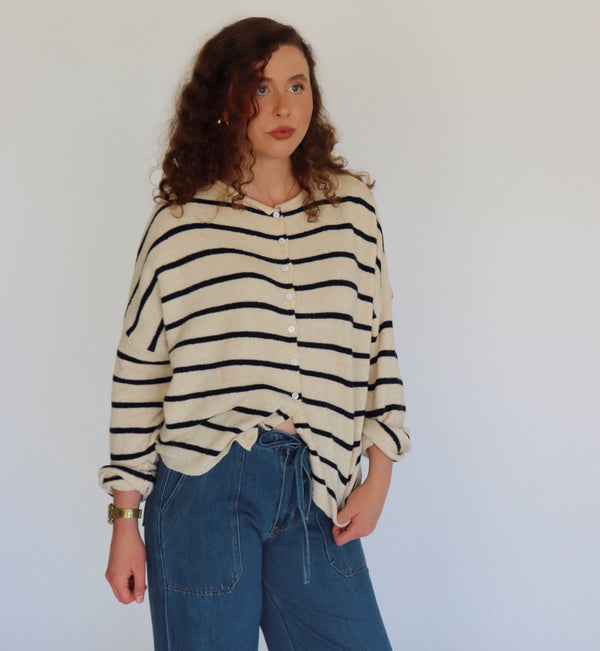 A woman with curly brown hair wearing a cream-colored knit cardigan with navy horizontal stripes and blue jeans, standing against a plain light gray background.