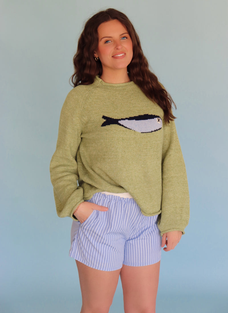 Woman wearing a green sweater with a fish design and blue shorts against a light blue background