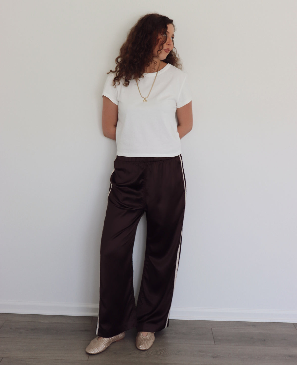 A woman stands against a white wall wearing dark brown wide-leg satin pants with white side stripes, a white short-sleeve t-shirt, and metallic beige shoes.