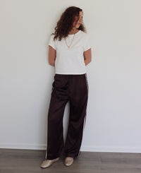 A woman stands against a white wall wearing dark brown wide-leg satin pants with white side stripes, a white short-sleeve t-shirt, and metallic beige shoes.