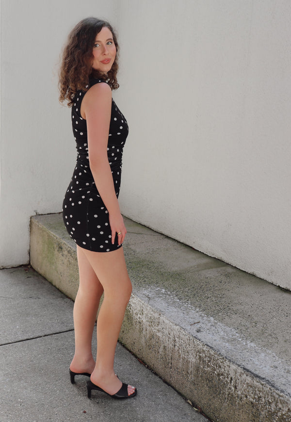 Woman in a black polka dot dress standing against a beige wall.