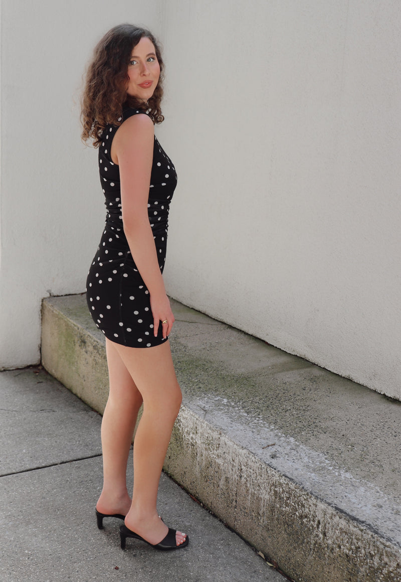 Woman in a black polka dot dress standing against a beige wall.