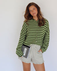 A woman modeling a green and dark green striped long-sleeve button-up cardigan with light beige ruffled shorts, holding a black and white patterned notebook against a plain white background.