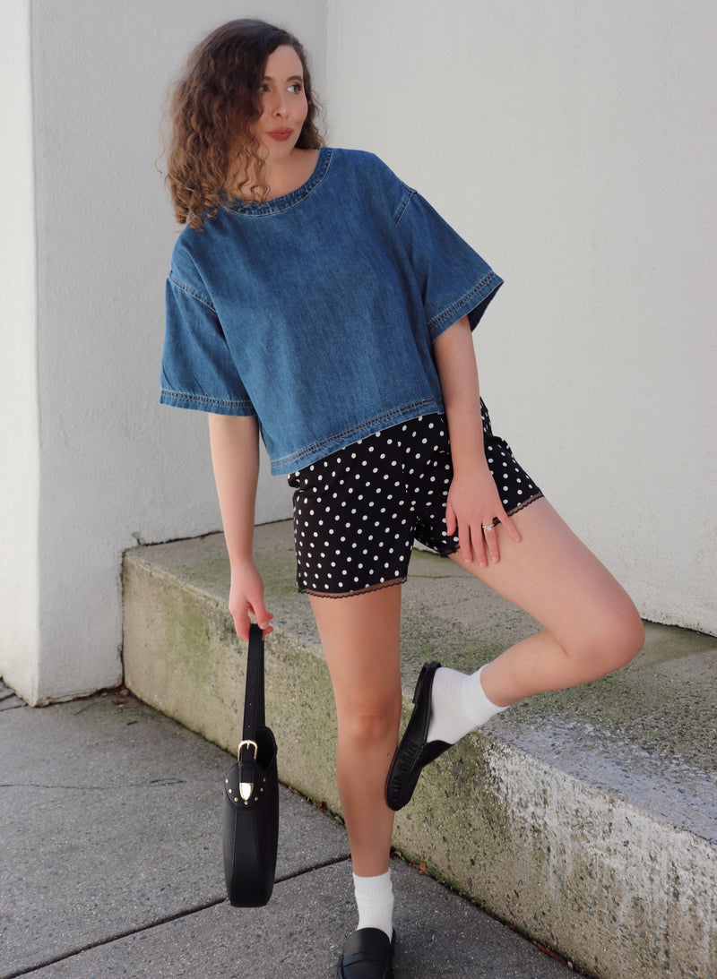 Woman wearing a denim shirt and polka dot shorts, standing against a beige wall.