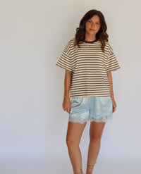 A woman modeling a beige and brown horizontally striped short-sleeve t-shirt with light blue satin shorts featuring a lace hem, standing against a plain white studio background.