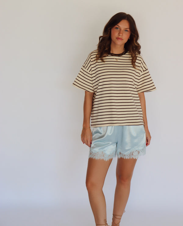 A woman modeling a beige and brown horizontally striped short-sleeve t-shirt with light blue satin shorts featuring a lace hem, standing against a plain white studio background.